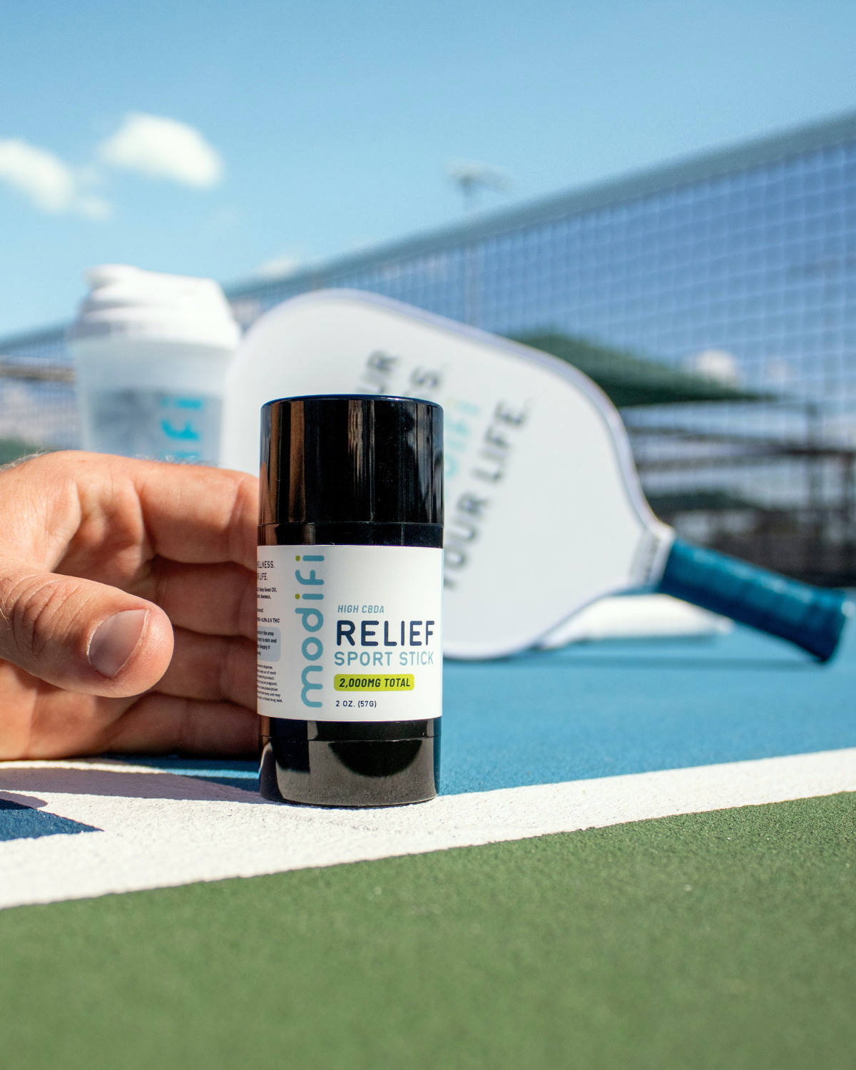 Hand holding a bottle labeled 'RELIEF SPORT STICK' on a tennis court.